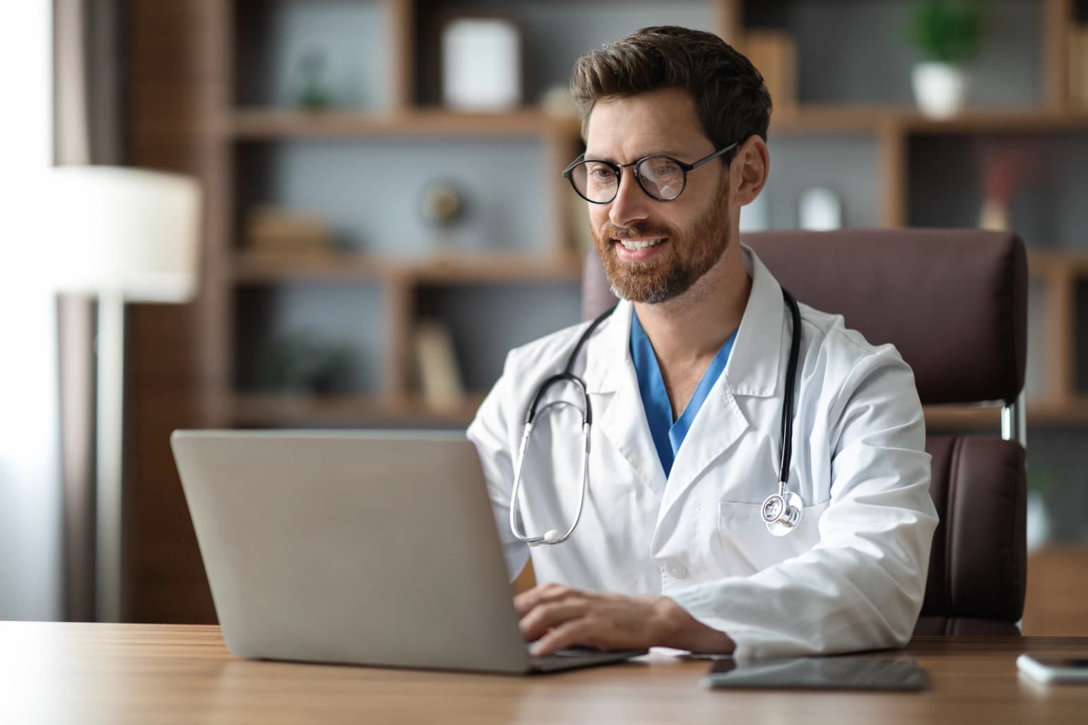 A physician in a white coat reviewing practice revenue on a laptop in a consulting room.