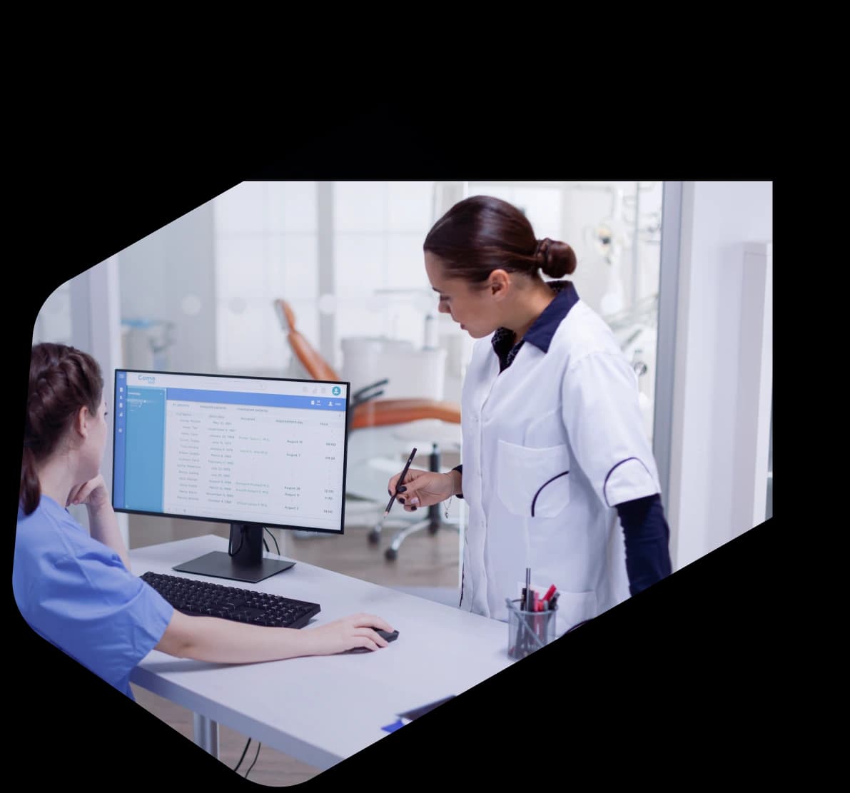 A medical coder in blue scrubs working through patient records at a clinic workstation while a colleague in a white coat reviews the screen beside her