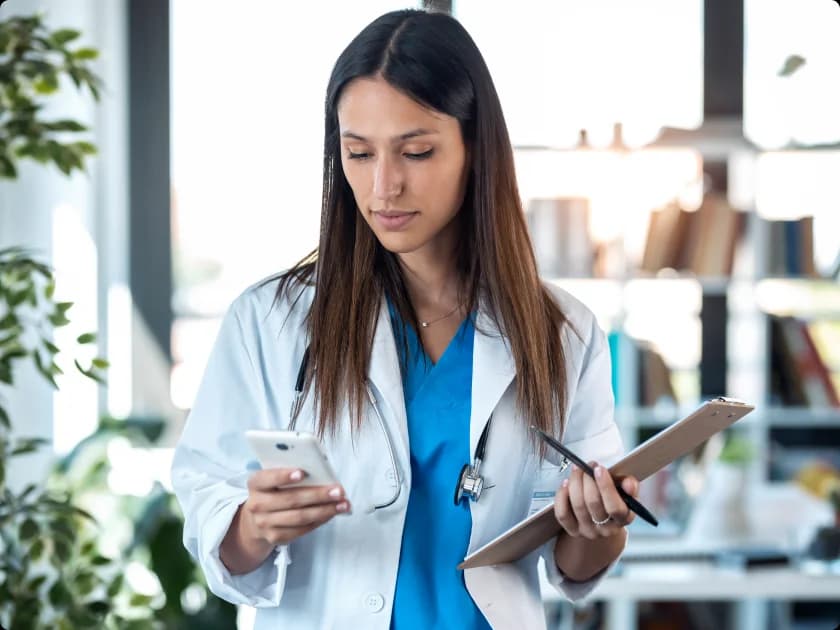 A clinician in a white coat over blue scrubs checking her phone while holding a clipboard and pen in a bright clinic office