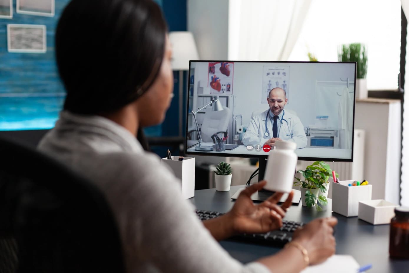 A woman seen from behind at her home desk on a video call with a doctor in a white coat and stethoscope on her monitor, holding up a white pill bottle, with a keyboard, a notepad and small potted plants on the desk