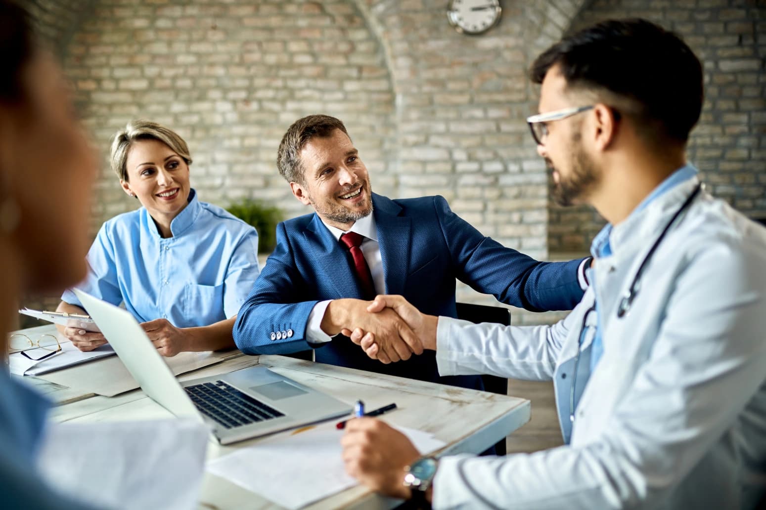 A practice administrator in a suit shaking hands with a physician across a desk, two clinical staff beside them with a laptop and notes
