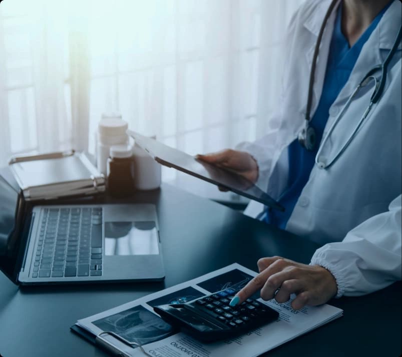 Physician in a white coat holding a tablet and working a calculator over a billing report beside a laptop