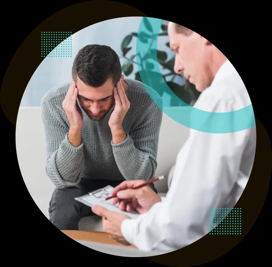 A clinician taking notes on a clipboard while a distressed patient sits with their head in their hands during a therapy session