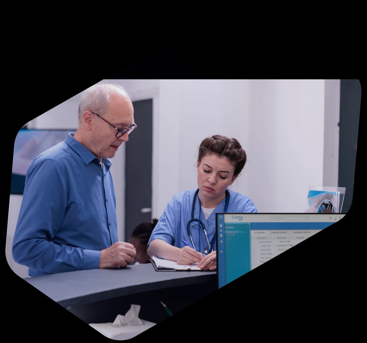 An older patient in a blue shirt at a clinic reception desk while a nurse in scrubs writes on a clipboard beside a screen of patient records