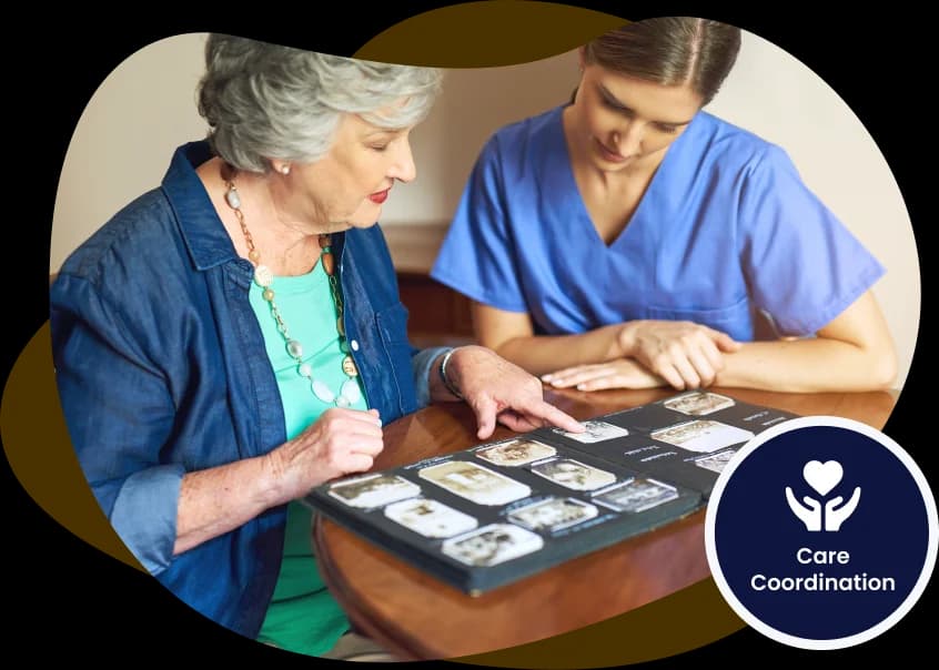 An older woman in a denim shirt and turquoise top at a wooden table with a carer in blue scrubs, the two of them looking through an open photo album she is pointing at, over a soft cream shape with a navy Care Coordination badge