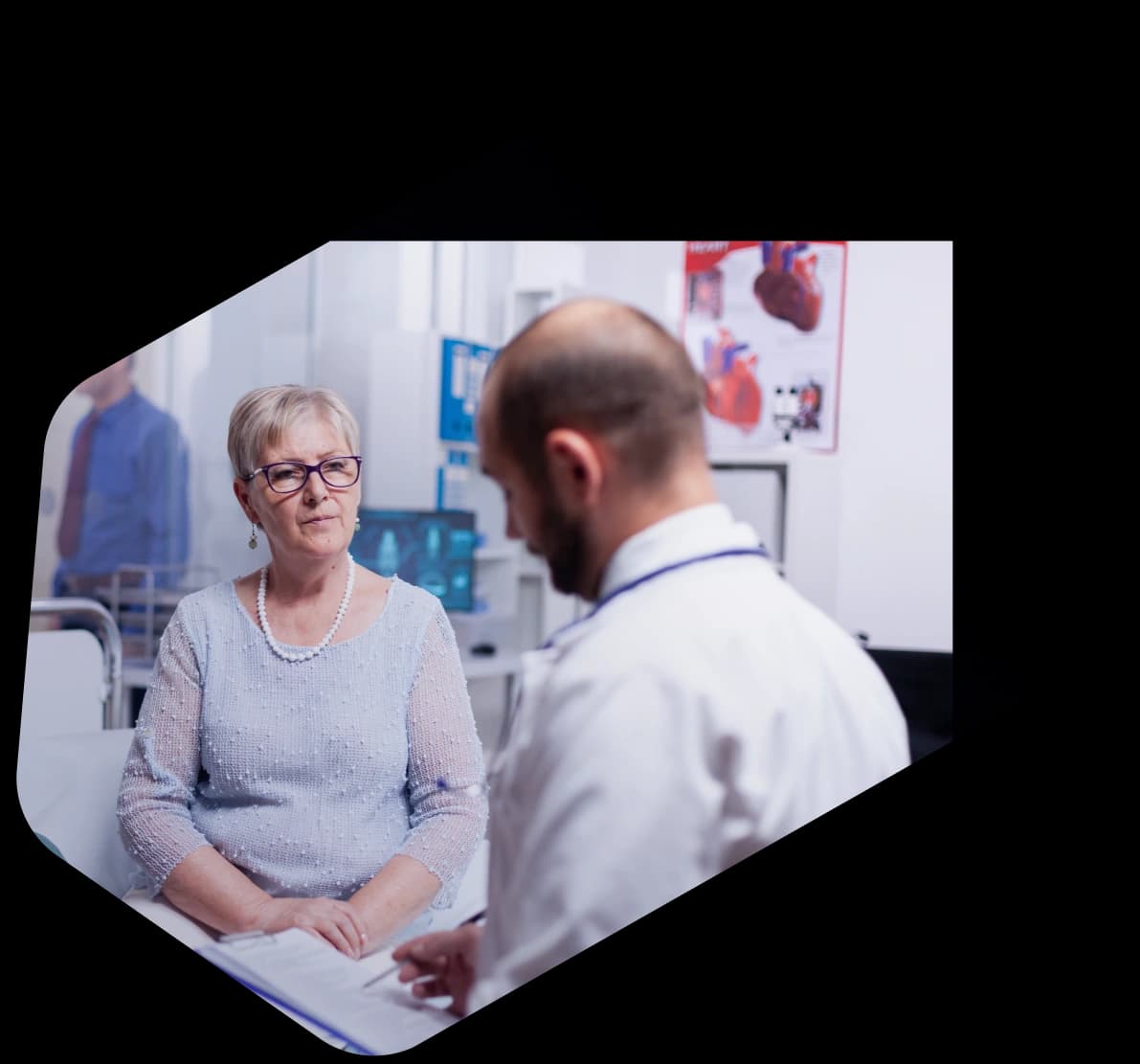 An older woman in glasses seated in a clinic exam room talking with a doctor in a white coat who is writing on a clipboard, a heart anatomy poster on the wall behind them