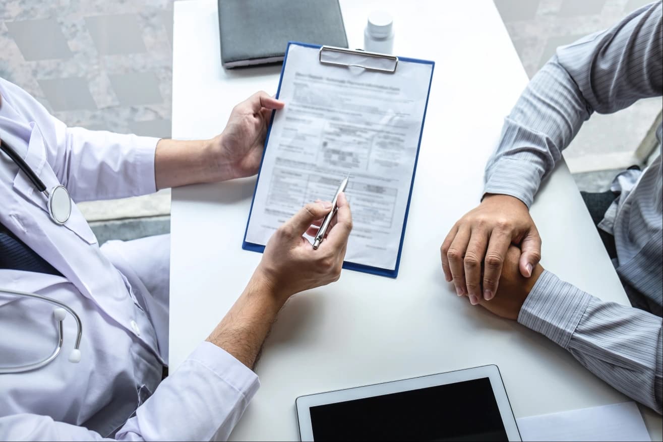 Overhead view of a clinician in a white coat with a stethoscope going through a claim form on a clipboard with a patient across the desk