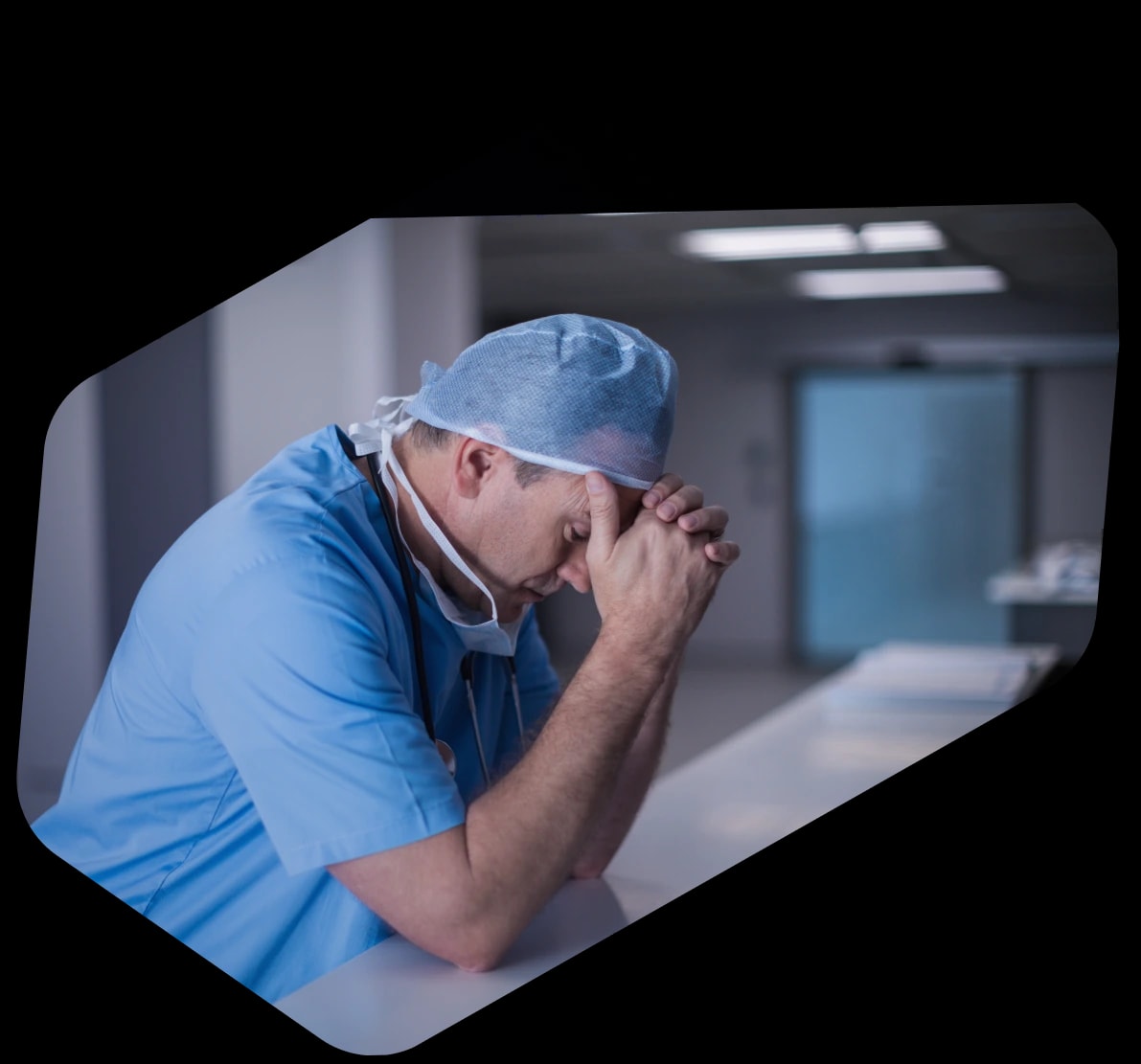 Doctor in blue scrubs and a surgical cap leaning on a hospital counter with his head resting on clasped hands