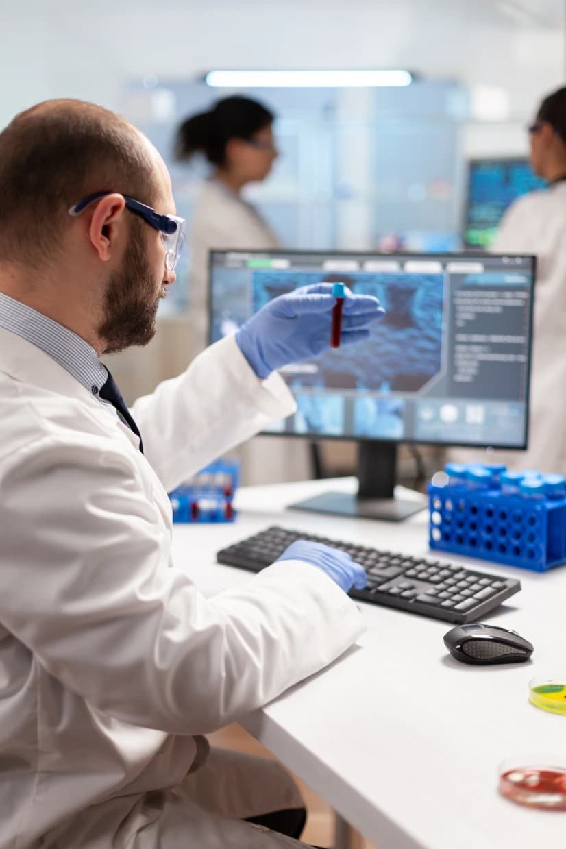 A laboratory scientist in a white coat and safety glasses holding a blood sample tube in front of a monitor showing analysis results, with colleagues working behind him