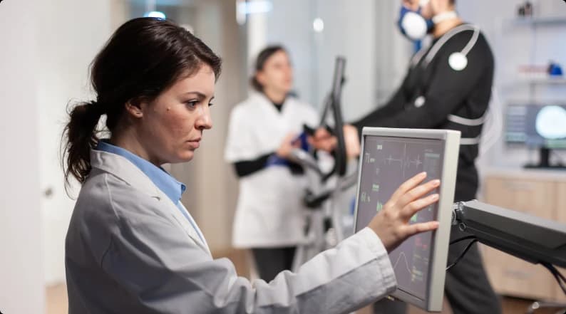 A scientist in a white coat adjusting a monitor showing waveform and vitals data while colleagues run a treadmill test on a masked patient behind her