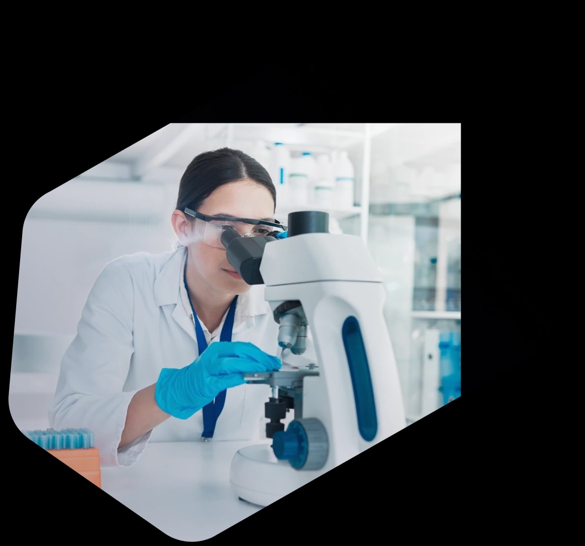 A laboratory scientist in a white coat, safety glasses and blue gloves adjusting a slide under a microscope at a lab bench, with specimen racks and reagent shelves behind her