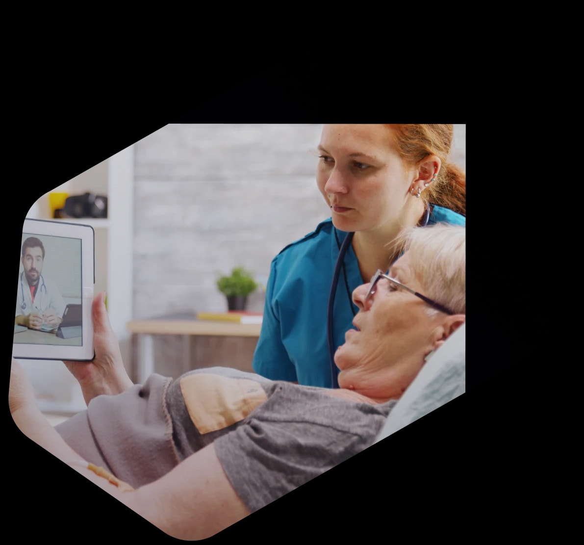 A home-care nurse leaning over an older patient resting in bed with a blood pressure cuff on her arm, both watching a doctor on a video call held up on a tablet