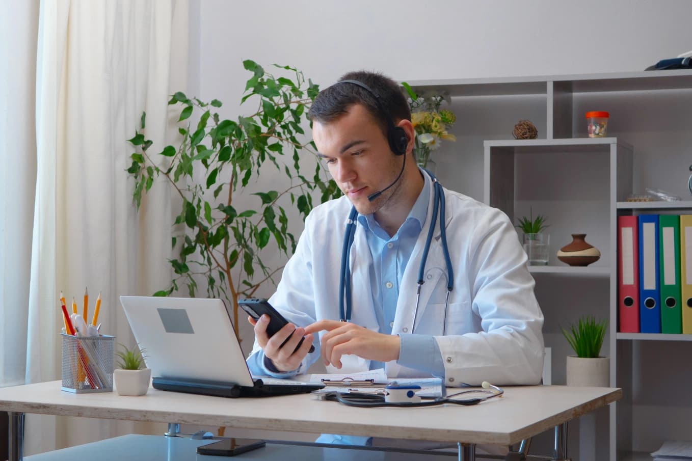 A clinician in a white coat with a stethoscope and a telephone headset at a desk, looking at a smartphone in his hand beside an open laptop, with a pen pot, small potted plants and shelves of ring binders behind him