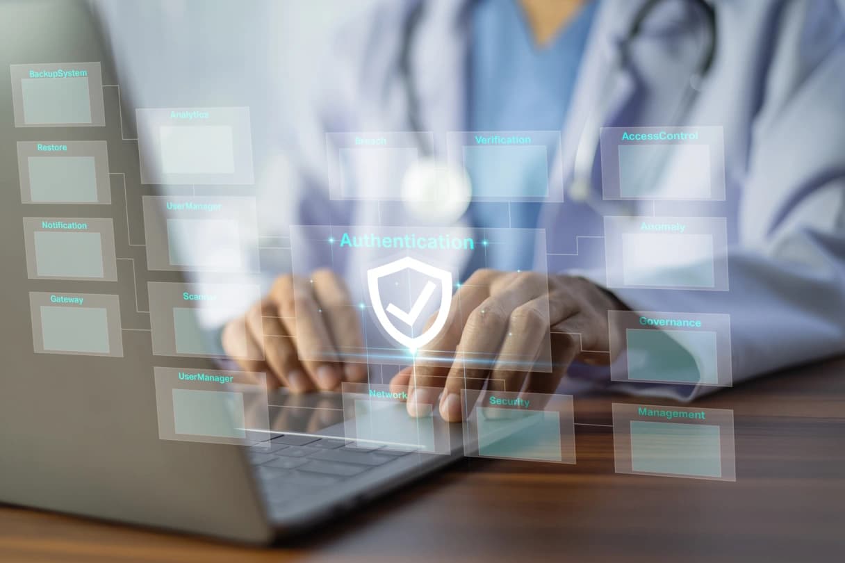 A clinician in a white coat and stethoscope typing on a laptop behind a translucent security dashboard, a white shield and tick at its centre labelled Authentication and surrounded by panels reading Access Control, Verification, Governance and Anomaly
